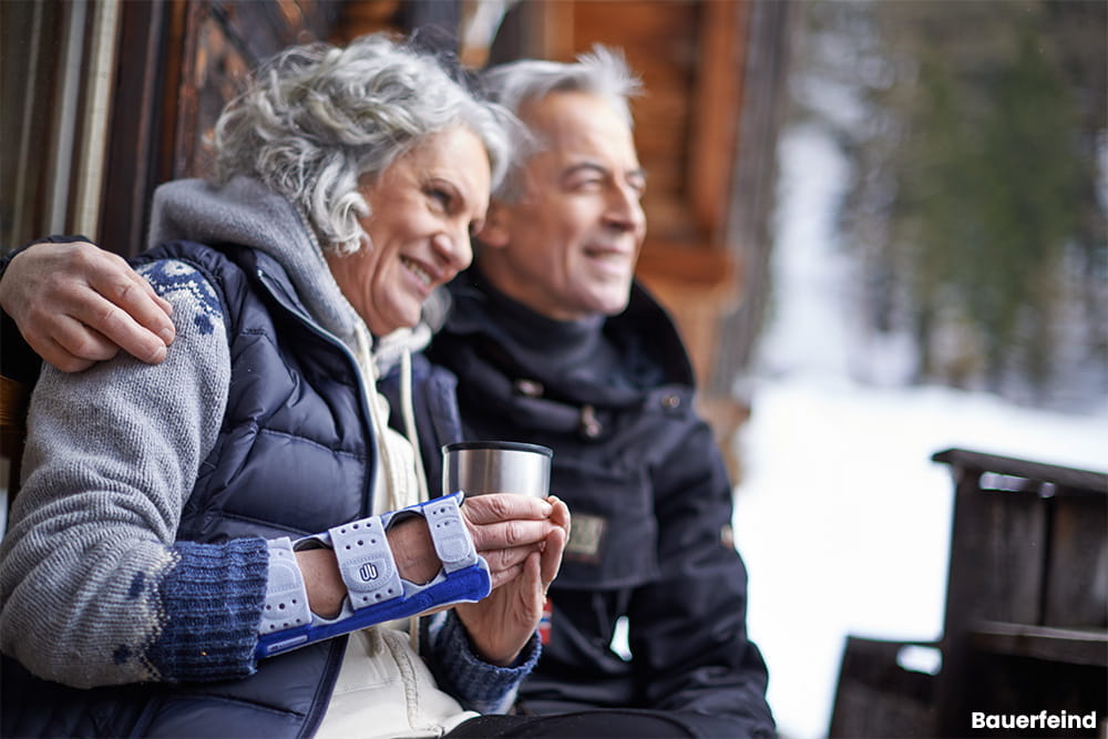Ein Paar sitzt vor einer Hütte im Schnee. Die Frau hält eine Tasse in der Hand und trägt dabei eine Handbandage.